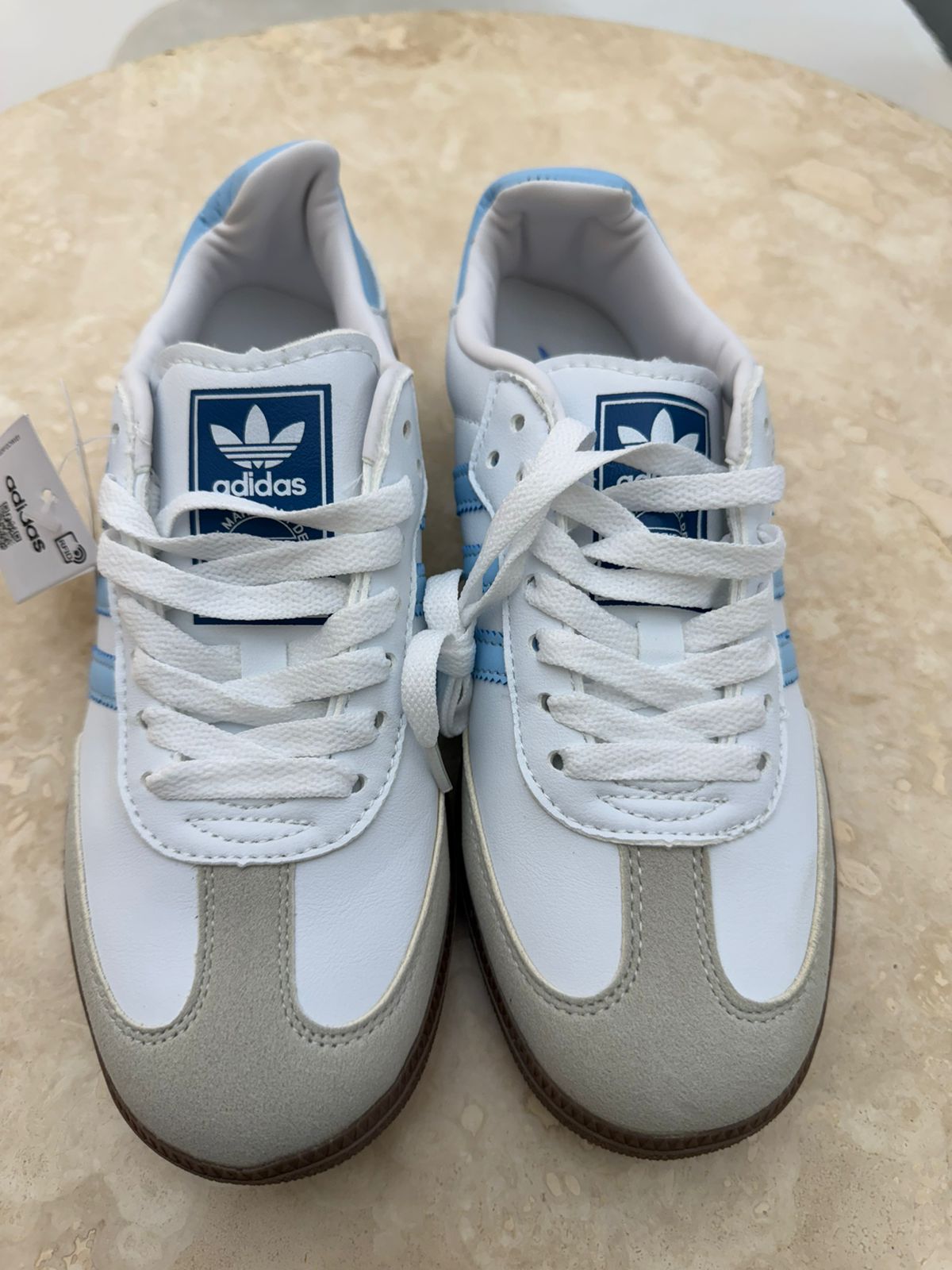 Adidas Samba OG sneakers feature a classic white leather upper with light blue three-stripes and a suede T-toe